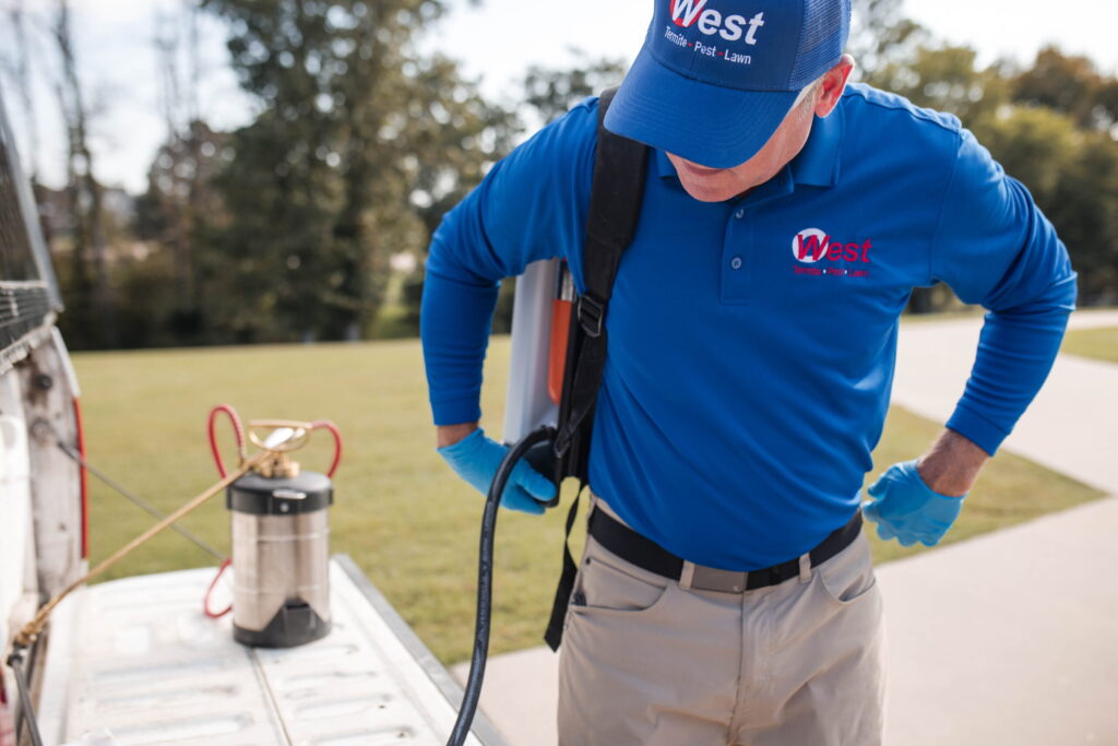 A person in a blue shirt and cap prepares pest control equipment from a truck bed, wearing gloves and a backpack sprayer, with grass and trees in the background.