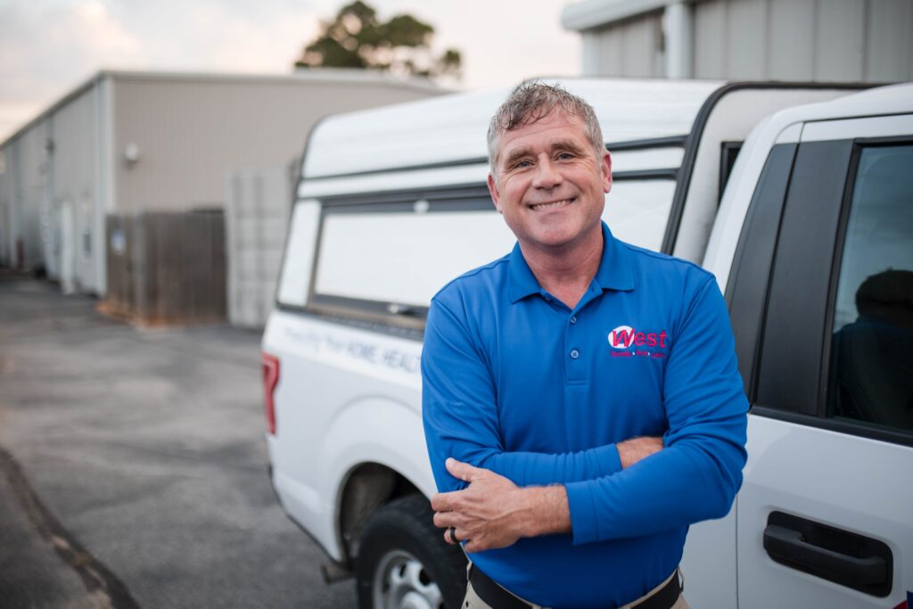 Smiling man in a blue polo shirt stands with arms crossed next to a white truck outside a building.