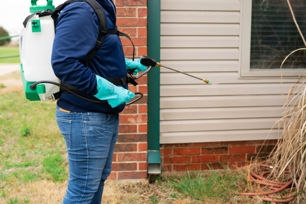 A person wearing gloves and a backpack sprayer applies pesticide or herbicide near the exterior wall of a house, next to dried grass and a garden hose.