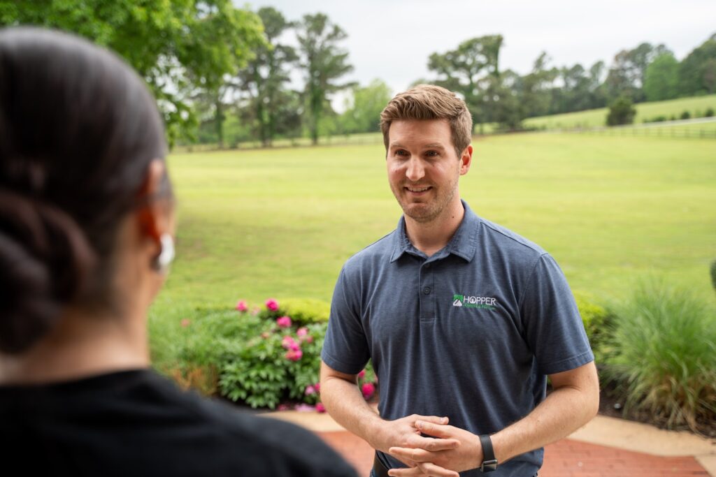 A man in a blue polo shirt talks to a woman outdoors near a garden, with a large grassy field and trees in the background on a cloudy day.