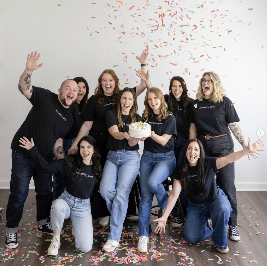 A group of young white presenting coworkers smile excitedly around two people with a cake and confetti