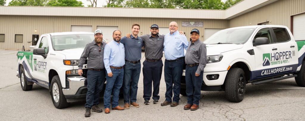 Six men wearing gray and blue shirts stand smiling between two white Hopper service trucks in a parking lot outside a beige building.
