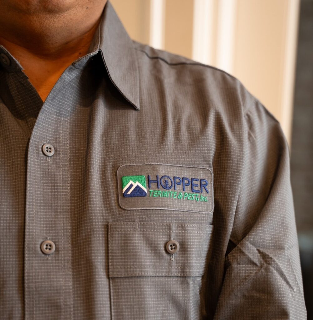 A person wearing a gray button-up shirt with a patch reading Hopper Termite & Pest, Inc. and a mountain logo on the front pocket.