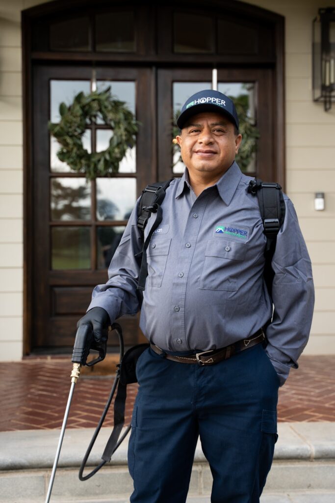 A pest control worker in uniform and gloves stands outside a house with holiday wreaths, holding spraying equipment.