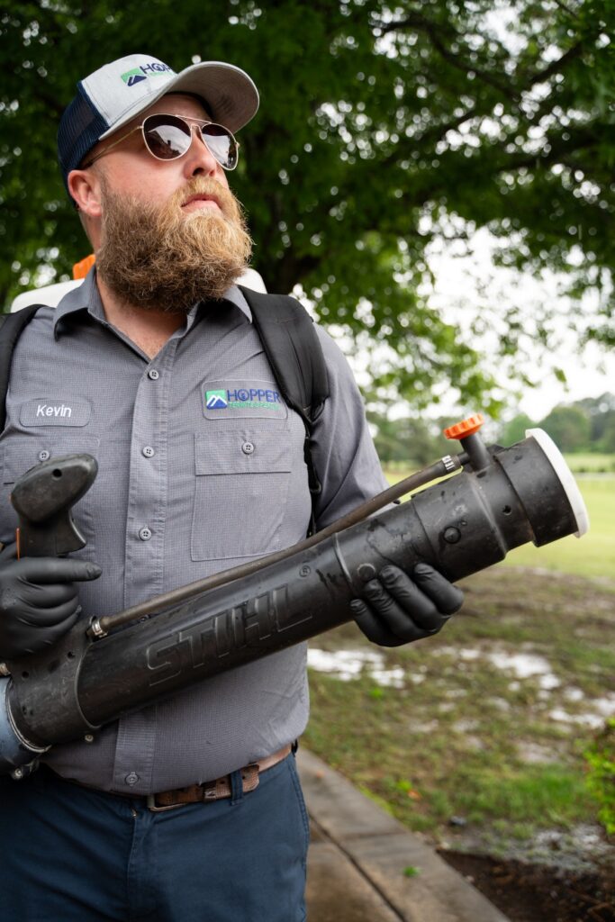 A bearded man in sunglasses and a gray Hopper work shirt holds a large black STIHL tool outdoors, standing near trees and grass after rain.