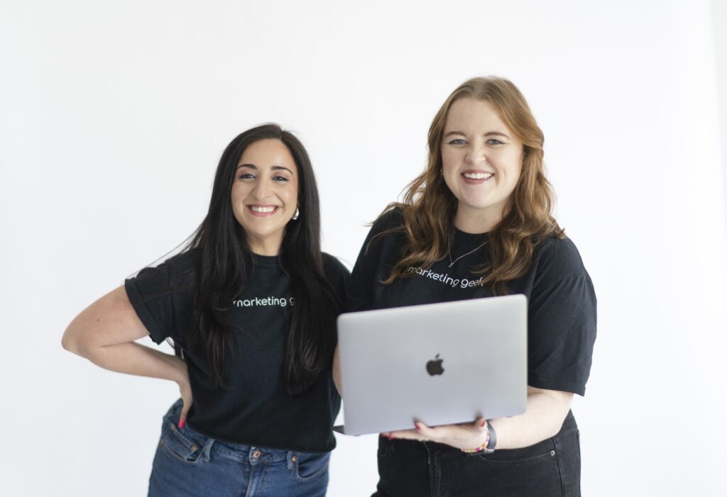 Two women smiling and standing together, both wearing black t-shirts and jeans, with one holding an open MacBook laptop. The background is plain white.