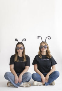 Two people sit cross-legged wearing marketing geek shirts, jeans, polka-dot bug-eye glasses, and curly antenna headbands, posing against a plain white background.