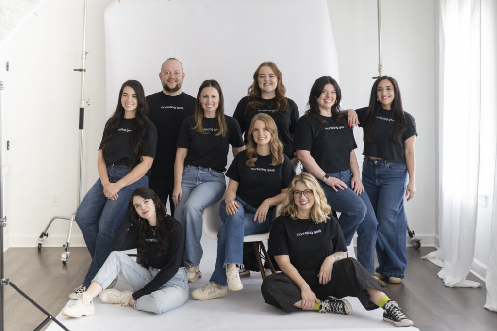 Nine people pose together in a bright studio, wearing black shirts and blue jeans. Some are standing, others are sitting or kneeling, all smiling at the camera. A white backdrop and photography equipment are visible.