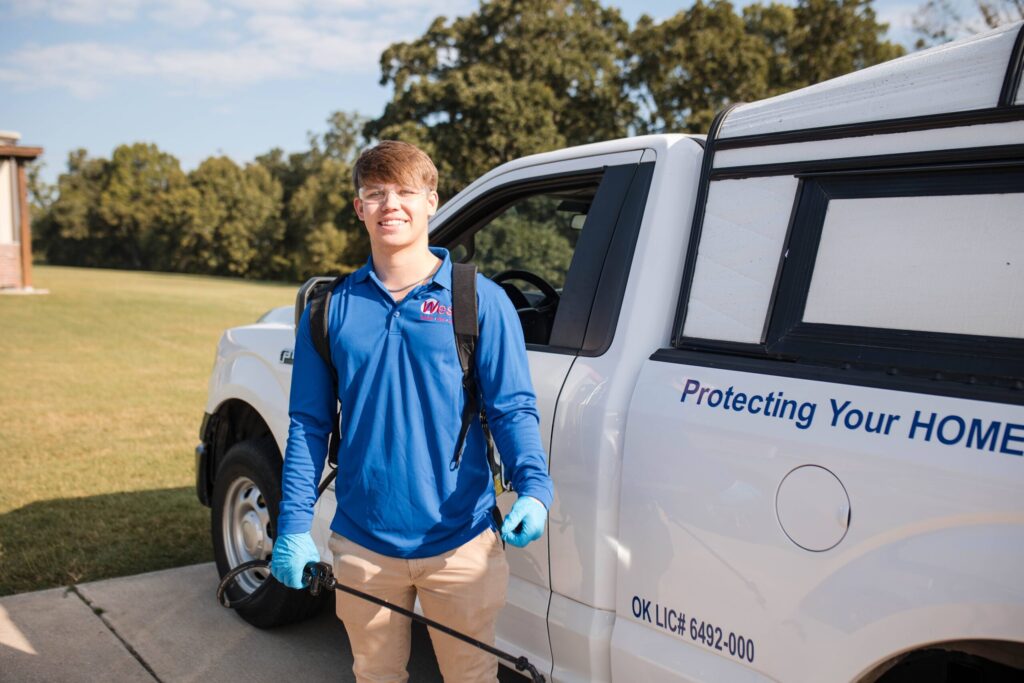 A person in blue uniform and gloves stands beside a white truck with Protecting Your Home on the door, holding pest control equipment, outside on a sunny day.
